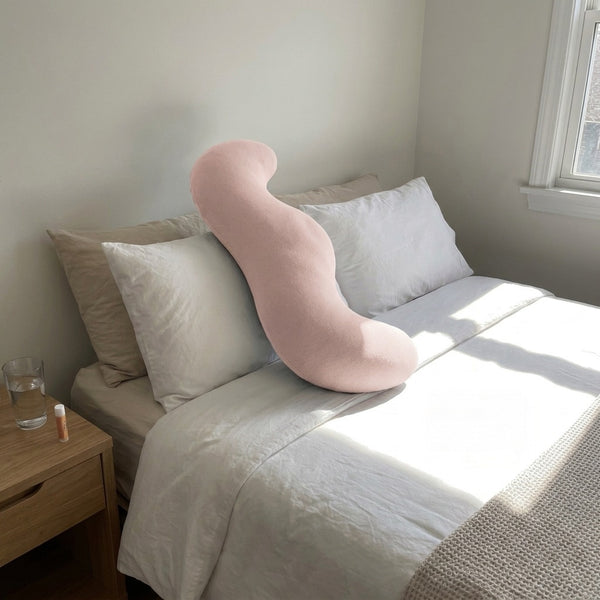 Blush pink Cuddle Curve pillow propped against the headboard in morning sunlight